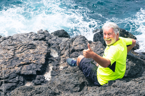 Happy senior man in outdoor excursion at sea sitting on the cliff looking at camera gesturing ok sign