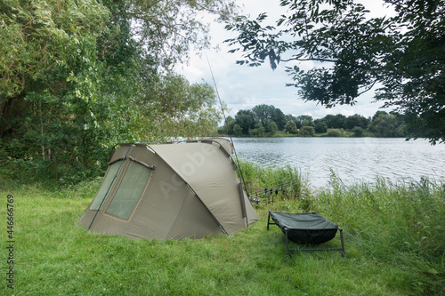 A green overnight fishing shelter on a lake side green grass bank.Several Rods and reels can be seen and a chair seat.