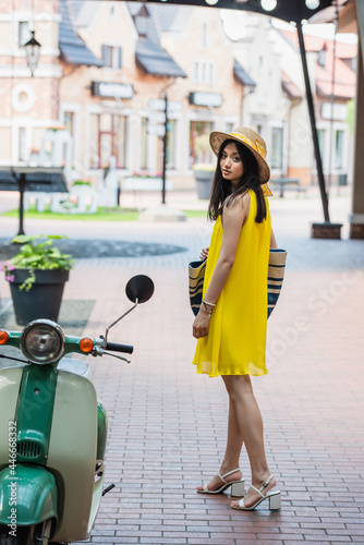 pretty asian woman in yellow sundress and sun hat looking at camera near scooter