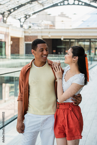stylish multiethnic couple looking at each other during summer walk in city