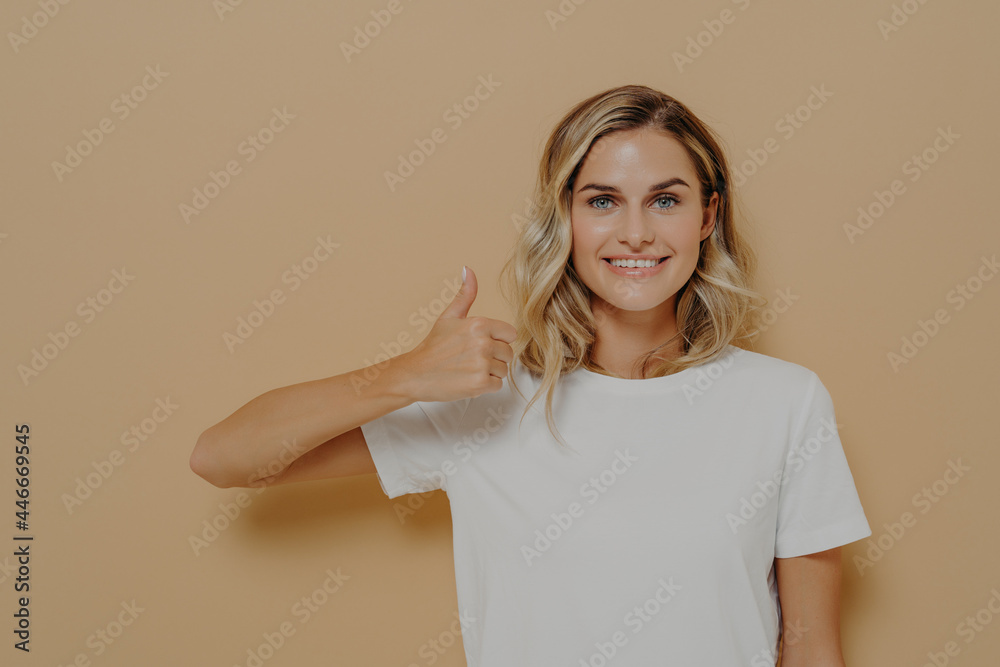 Happy young blonde female wearing white casual tshirt making thumbs up gesture and smiling cheerfully Happy young blonde female wearing white casual tshirt making thumbs up gesture and smiling cheerfully