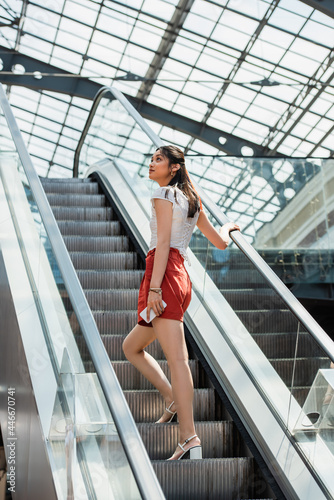 young asian woman holding smartphone while looking away on escalator