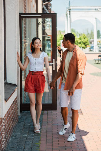 stylish multiethnic couple looking at each other and holding hands near building on street