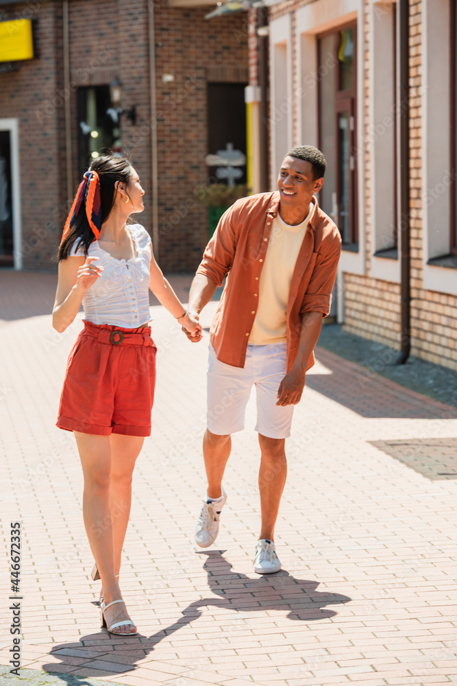 excited african american man holding hands with asian woman on urban street