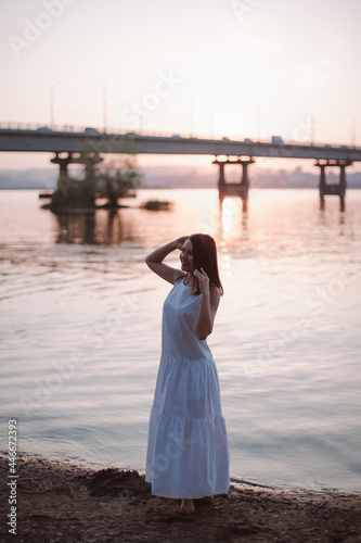 Women posing at sunset. Full-length portrait of a beautiful, young woman posing at sunset on the beach with a view of the bridge over the river and the silhouette of the city. 