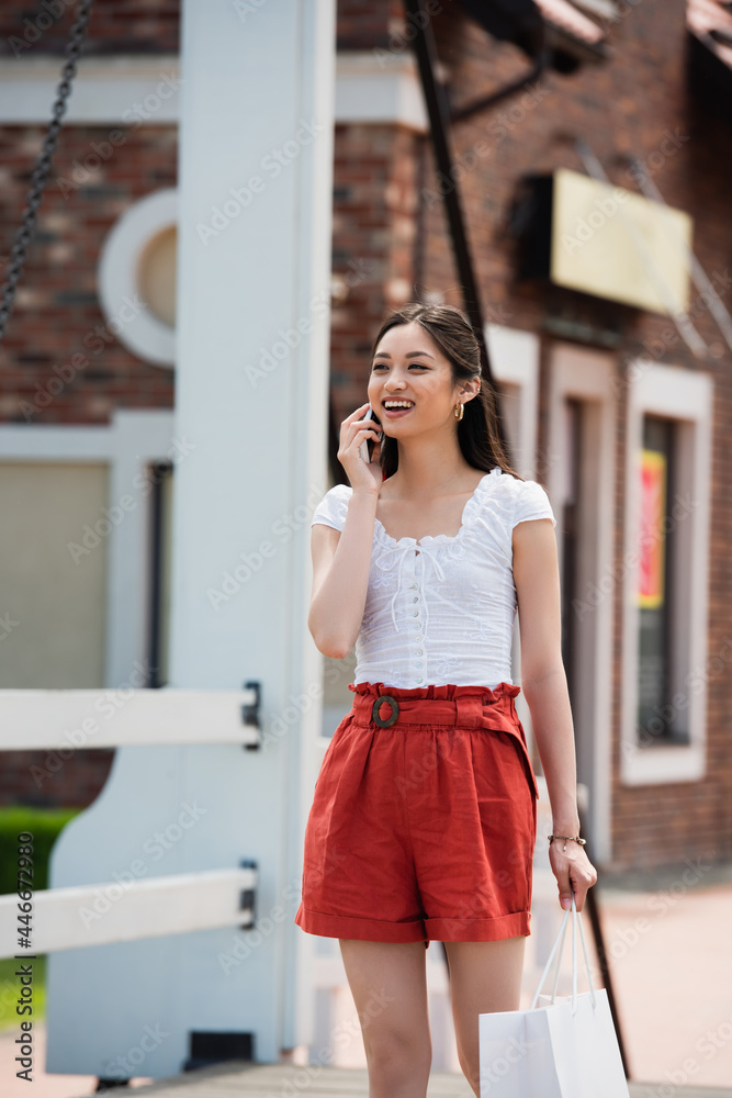 cheerful asian woman calling on smartphone while walking with shopping bag on bridge