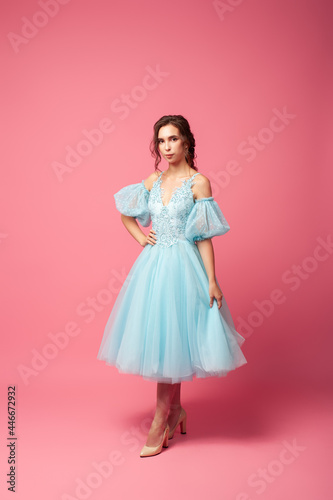 A young woman in an airy dress with sleeves and beige shoes poses against an isolated background. A beautiful brunette in a blue ball gown and evening hairstyle holds an airy skirt in her hand. 