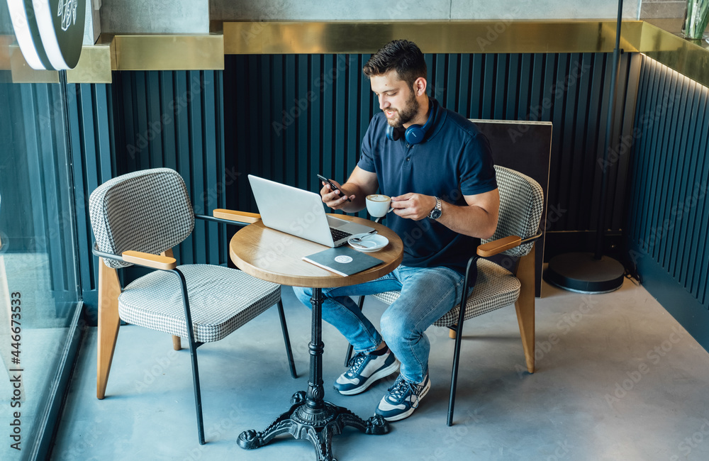 Casual Businessman with Headphones Working on Laptop and Mobile Phone in a Cafe.
Young business man using his phone while working on laptop and drinking coffee in a restaurant table.