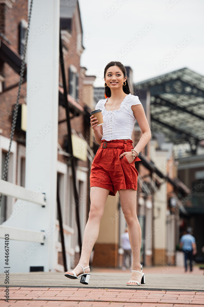 joyful asian woman holding coffee to go while walking on urban street with hand in pocket