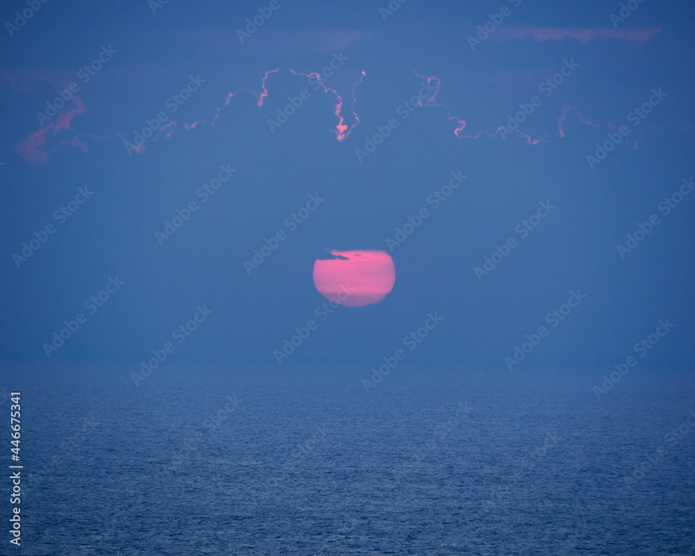 Fototapeta premium sunset, a magenta pink sun setting over the horizon with a calm sea and a sky with few clouds