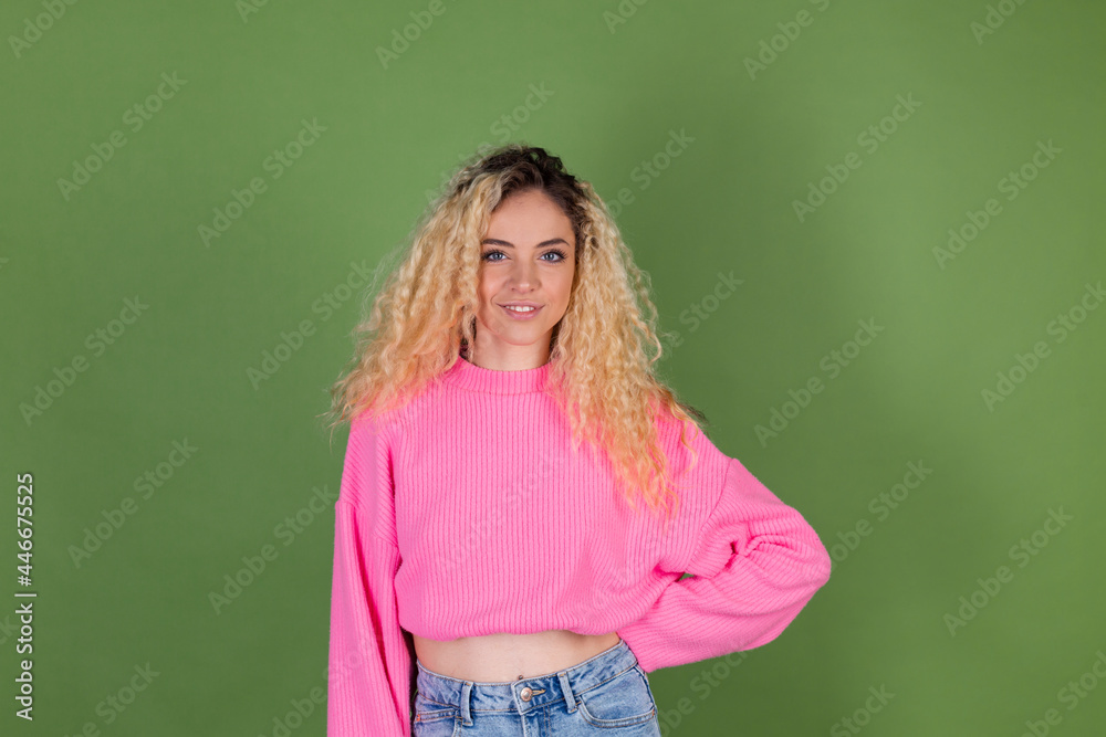 Young blonde woman with long curly hair in pink sweater on green background positive smiling look to camera Young blonde woman with long curly hair in pink sweater on green background positive smiling look to camera