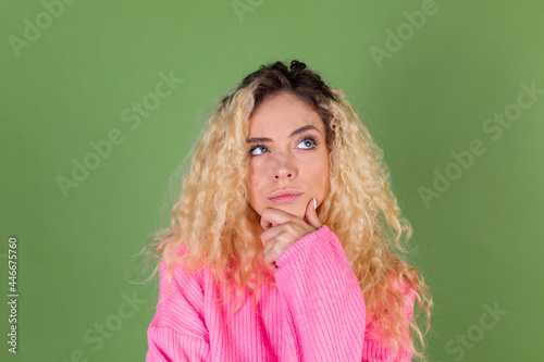 Young blonde woman with long curly hair in pink sweater on green background thoughtful hand on chin look up