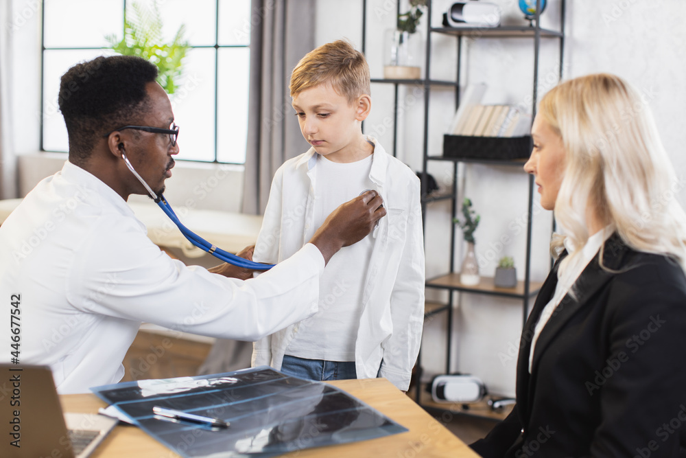 Fototapeta premium Experienced black pediatrician using stethoscope for examining heartbeat of little boy. Pretty caucasian mother sitting at table while doctor doing checkup for her son. Health care concept.