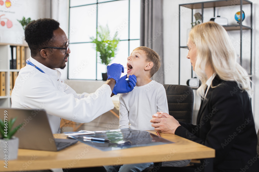 Caucasian woman and her lovely son having medical appointment at modern clinic. African pediatrician examining throat of little patient at cabinet. Concept of regular checkup for children.