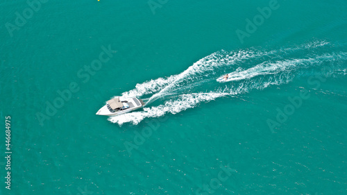 Aerial drone photo of woman practicing high speed water ski towed by large speed boat in emerald tropical lake
