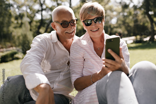 Charming lady with short hairstyle in sunglasses and pink blouse looking into smartphone and smiling with grey haired man in white shirt in park.. © Look!