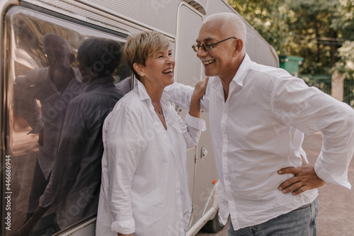 Cheerful short haired woman in light cool blouse laughing with man with grey hair and mustache in white shirt and eyeglasses outdoors..