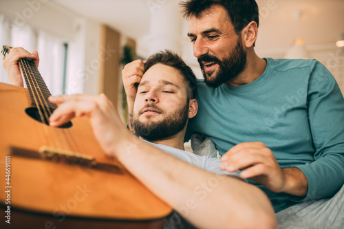 Happy gay couple in love having romantic times at home. Man is playing guitar to his beloved boyfriend.