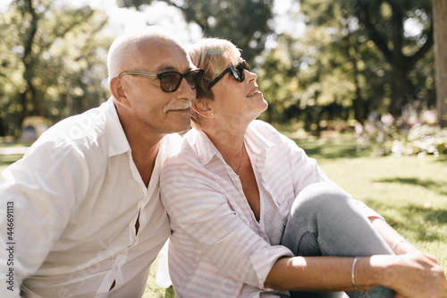 Grey haired man with mustache in sunglasses and light shirt smiling and sitting on grass with woman with short hair in pink blouse outdoor.. © Look!
