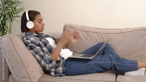 A woman in jeans with headphones using a silver laptop, lying on a sofa in a bright apartment. The concept of working and learning online.