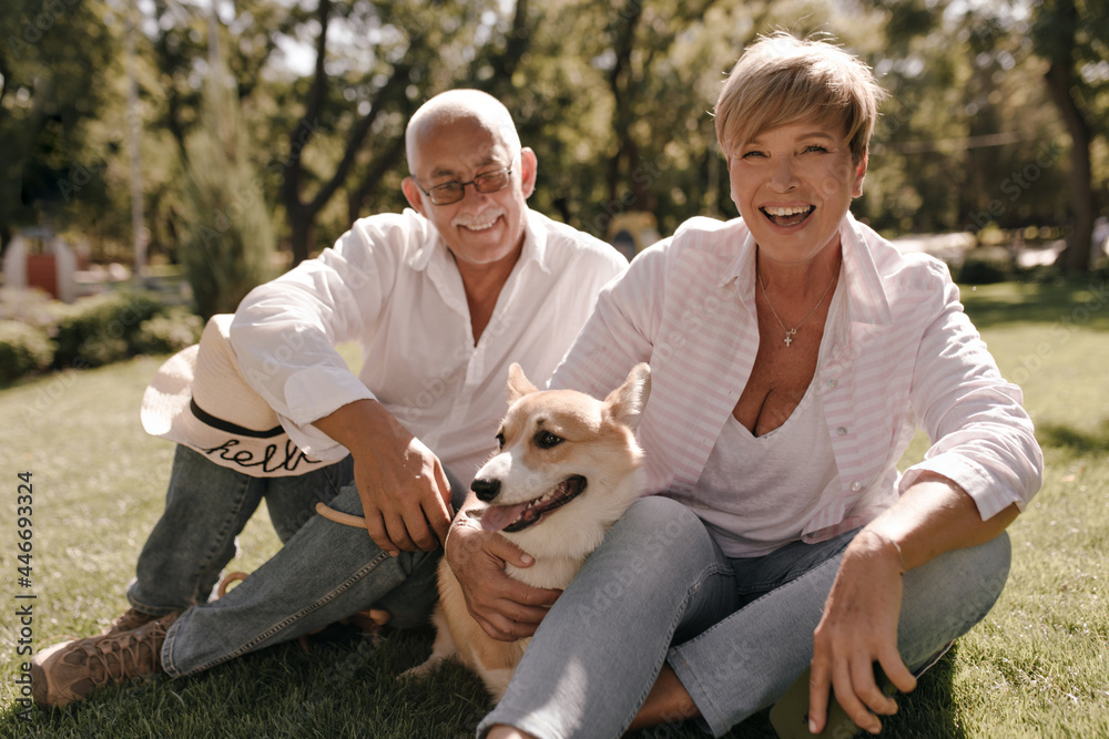 Joyful woman with blonde trendy hairstyle in pink shirt and jeans laughing, hugging corgi and sitting with grey haired man in eyeglasses in park..