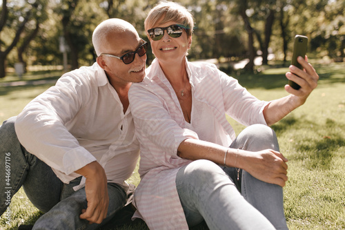 Happy woman with blonde hair in pink clothes and sunglasses smiling, sitting on grass and making photo with grey haired man in white shirt in park.. © Look!