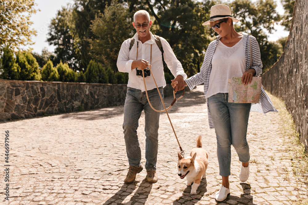 Obraz premium Wonderful woman in hat, white t-shirt, striped blouse and jeans holding card and hand grey haired man in light clothes with dog in park..