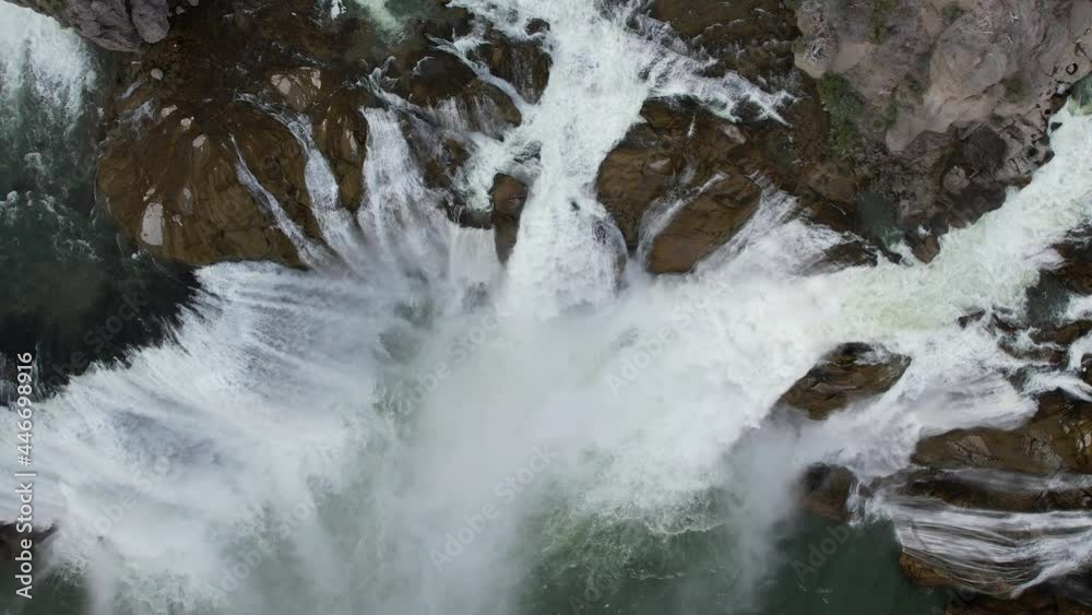 Aerial birds eye view of powerful waterfall flowing over rocks in slow motion while drone slowly descends towards cascading water