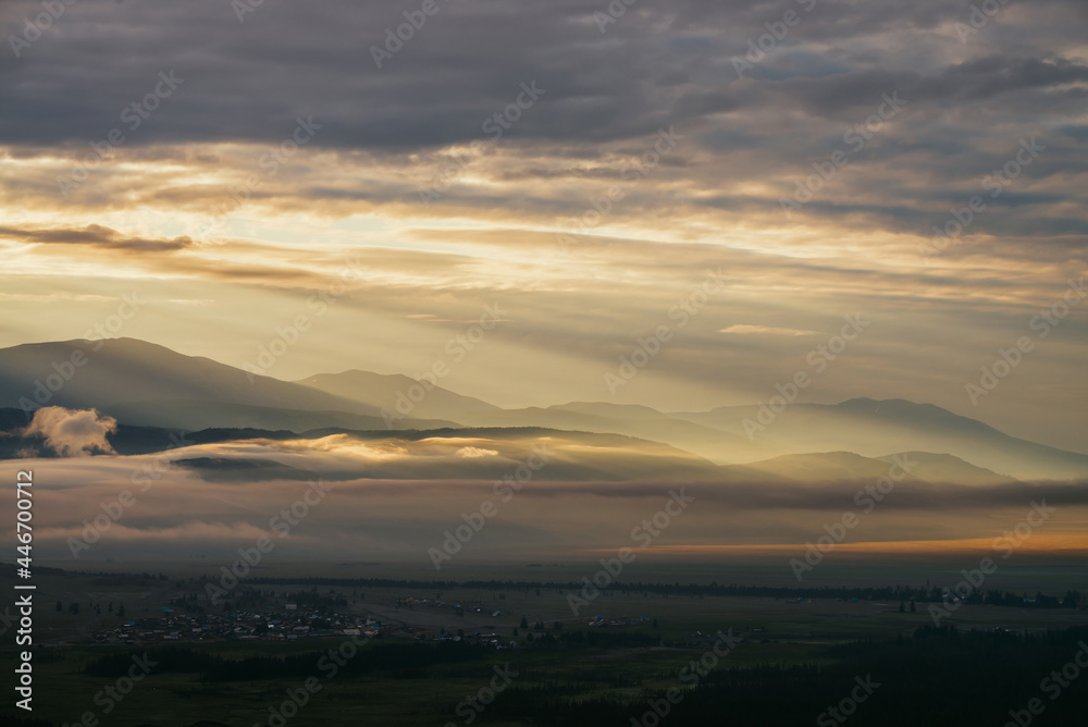 Fototapeta premium Scenic mountain landscape with golden low clouds above village among mountains silhouettes under dawn cloudy sky. Atmospheric alpine scenery of countryside in low clouds in sundown illuminating color.