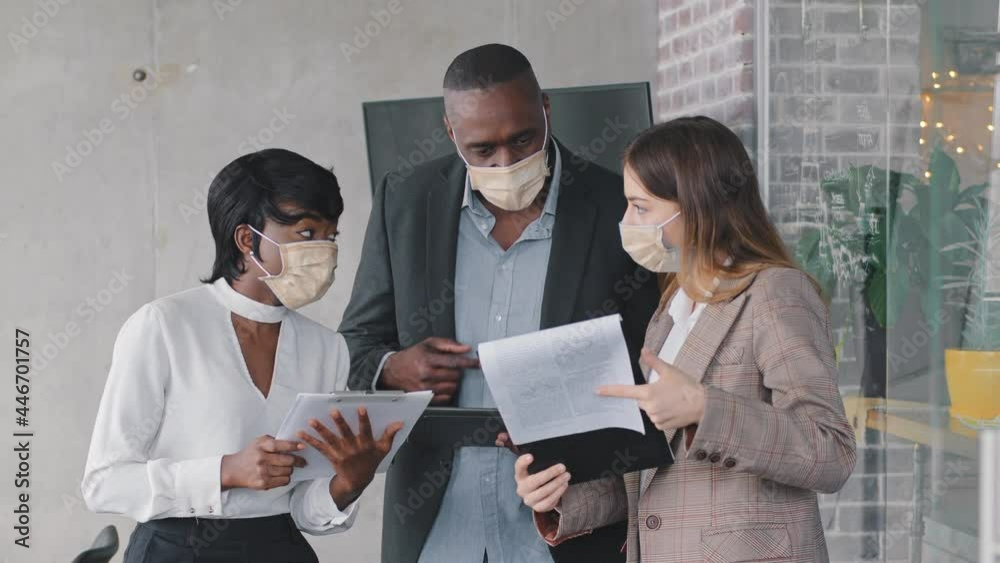 Multiracial business team adult mature african man boss, afro black woman manager and caucasian businesswoman standing in office wearing protective medical masks holding documents papers checking data Multiracial business team adult mature african man boss, afro black woman manager and caucasian businesswoman standing in office wearing protective medical masks holding documents papers checking data