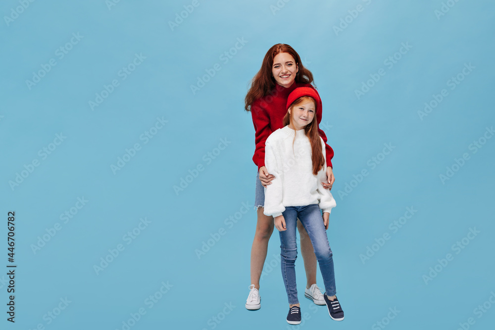 Lovely young girl in red hat and modern jeans smiling and posing with elder sister in white cool sneakers on isolated background.. Lovely young girl in red hat and modern jeans smiling and posing with elder sister in white cool sneakers on isolated background..