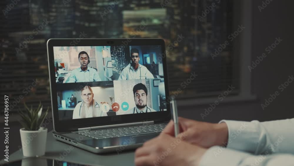 Computer monitor with video conference of multiracial doctors' leaders. Hands writing notes in front. Computer monitor with video conference of multiracial doctors' leaders. Hands writing notes in front.