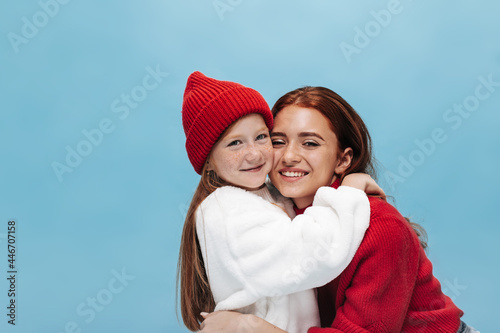 Photo of two pretty sisters in modern outfit. Young girl in white clothes hugs smiling lady with brunette hair on isolated background..