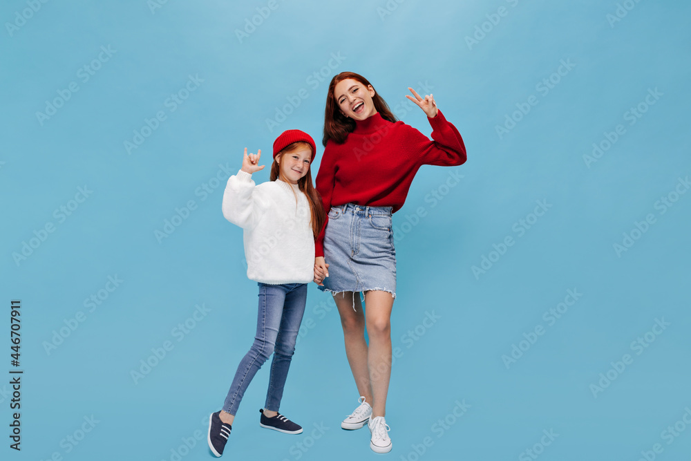 Wonderful woman in red shirt, denim skirt and sneakers shows peace signs and holds hand her young sister with ginger hair in jeans on blue background..