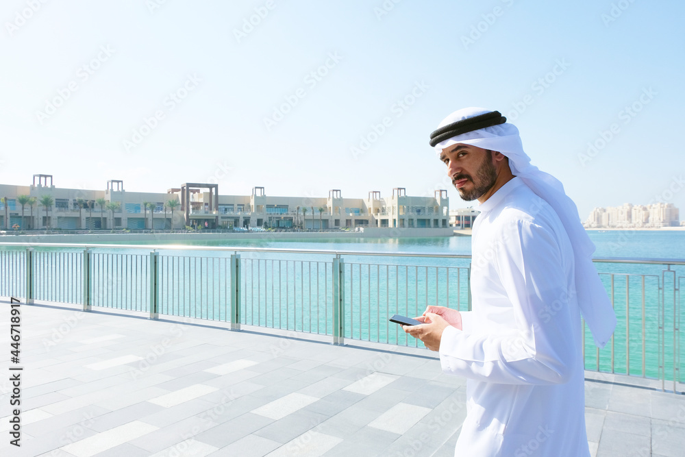 Arabic man using mobile smart phone. Emirati on Kandura holding a device while checking online websites, eCommerce or blogs Arabic man using mobile smart phone. Emirati on Kandura holding a device while checking online websites, eCommerce or blogs