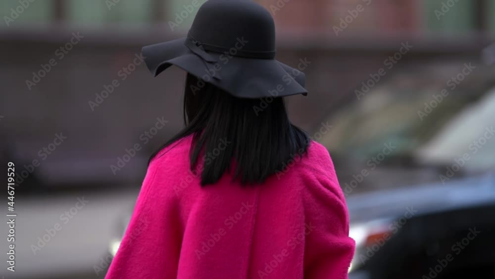 a brunette in a black hat and a bright pink coat stands against the background of a blurred city street and a passing car. rear view. the camera is moving a brunette in a black hat and a bright pink coat stands against the background of a blurred city street and a passing car. rear view. the camera is moving