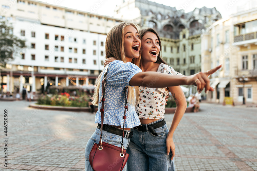 Portrait of excited surprised girlfriends in stylish jeans and floral blouses pose outside. Blonde girl in blue top points into distance.