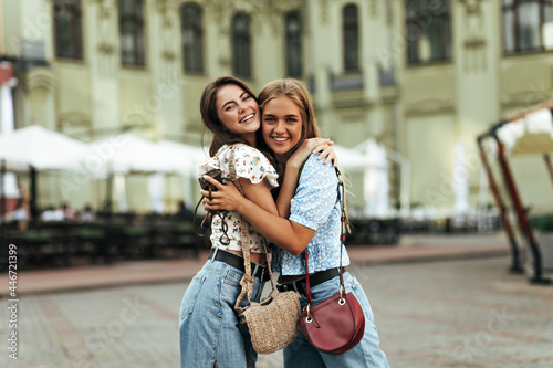 Joyful brunette and young blonde women in stylish denim pants and colorful blouses rejoice, have fun, smile widely outdoors.