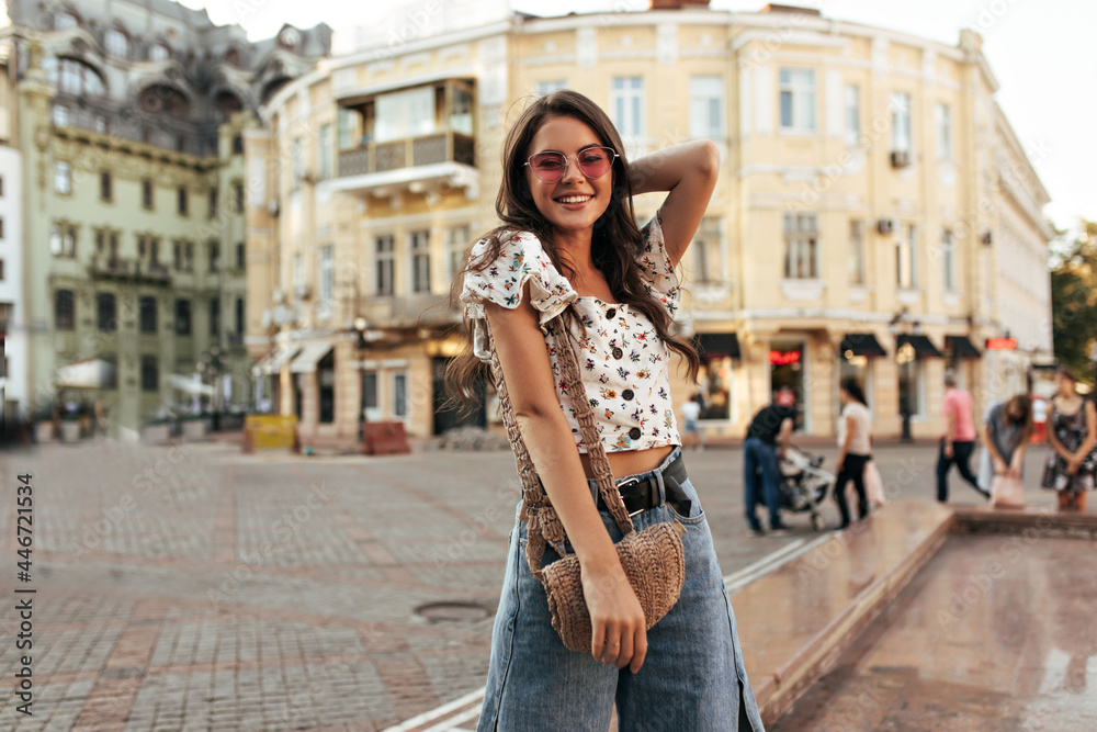 Happy young brunette woman in loose stylish denim pants and cropped floral top smiles widely. Curly girl in pink sunglasses poses in good mood outdoors.
