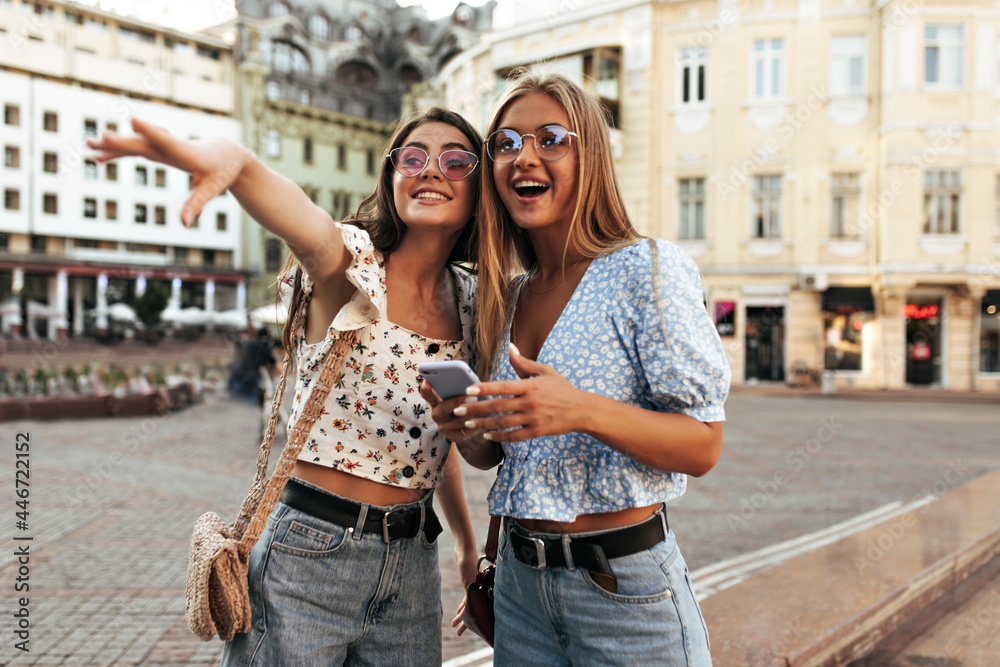 Cheerful attractive women in stylish summer outfits look into distance with surprise. Excited blonde and brunette girls in colorful sunglasses pose outdoors and smile.