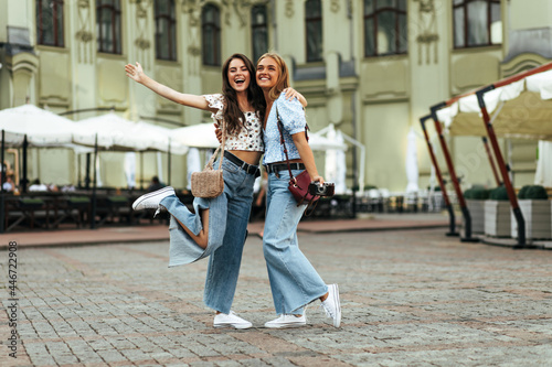 Charming pretty girls in stylish denim pants and floral tops hug and  rejoice. Attractive lady in jeans and white blouse smiles and rises arm and  leg outside. Stock Photo