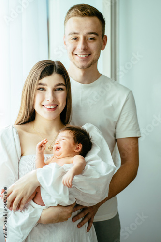 Vertical portrait of happy family of three with newborn baby. Mother and father look at camera, smile, holding sweet adorable healthy newborn child carefully on arms. All dressed in white clothes.