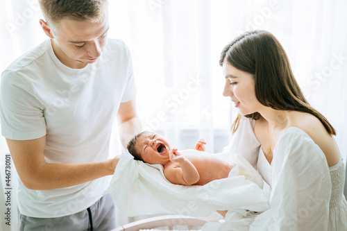 Relationship with husband after childbirth concept. Adorable cute baby new born girl shouting on parents hands. Crying child in sunny bedroom. Young family try to calming baby in light cozy bedroom..