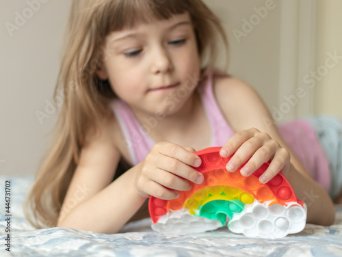 Little girl lies on the bed and plays with pop it in the shape of a rainbow, squashing bubbles with her fingers