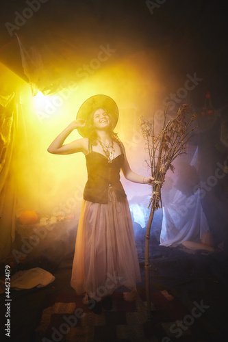 Young slim woman looking like witch having fun on Halloween in a dark room with yellow light and smoke. Carnival concept and Halloween party