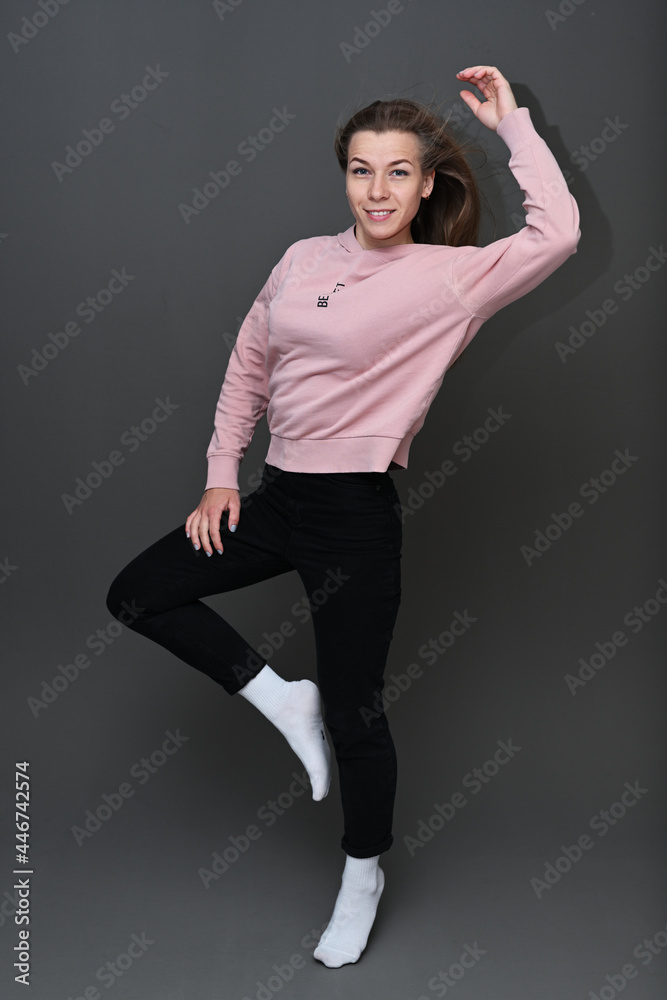 Full length portrait of a young caucasian girl jumping with a smile and fluttering hair posing in a pink sweater and black trousers on a dark gray background