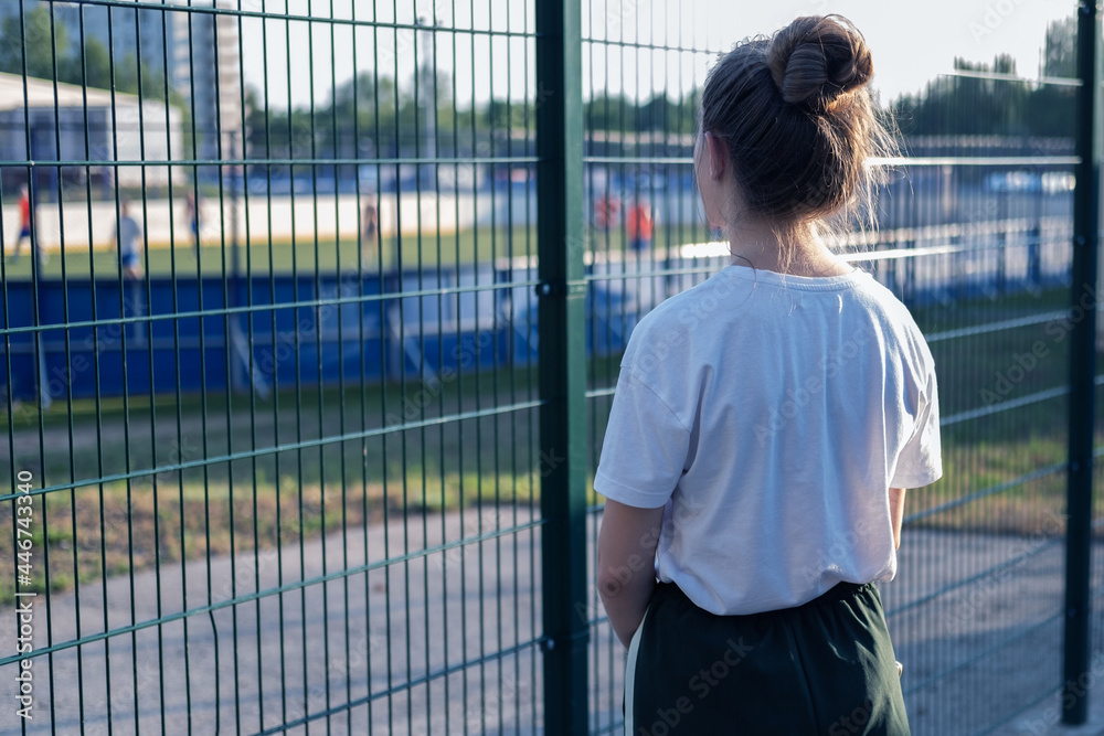 Fototapeta premium A girl stands and looks through the fence at the athletes playing at the stadium. The girl wants, but cannot play sports.