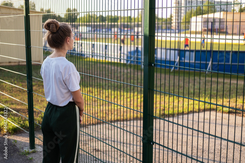 A girl stands and looks through the fence at the athletes playing at the stadium. The girl imagines how she will go in for sports and play at the stadium.