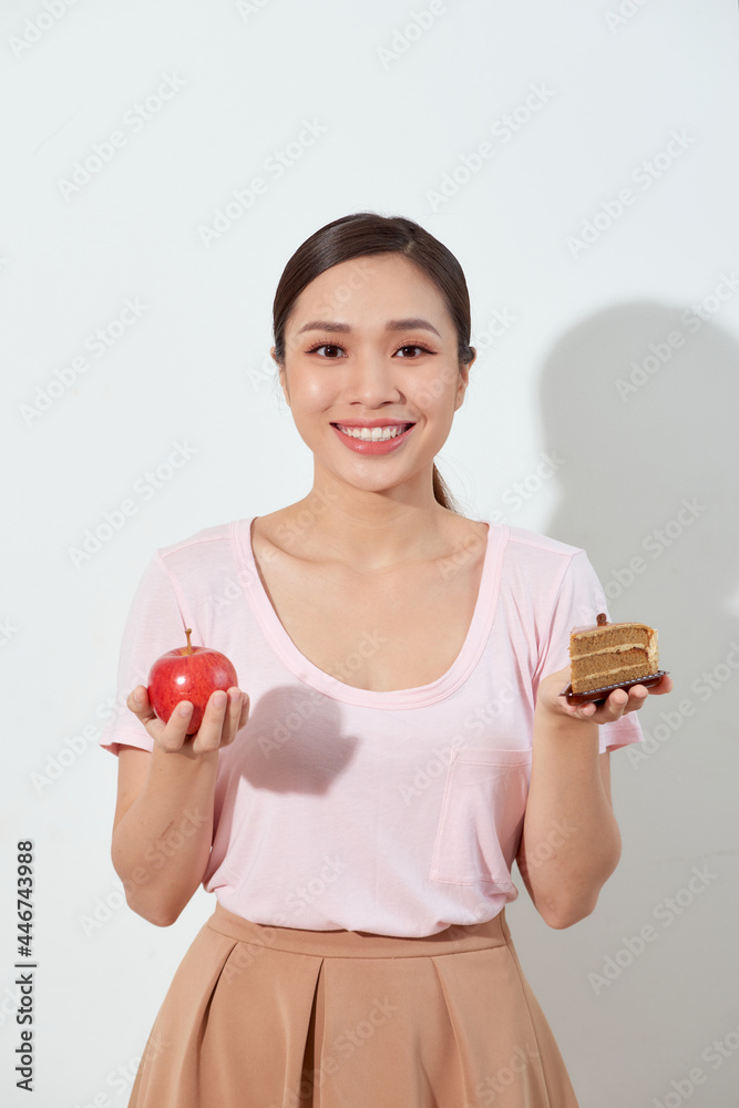 Portrait of a confused young asian woman choosing between cakes and apple, isolated on a white background. Portrait of a confused young asian woman choosing between cakes and apple, isolated on a white background.