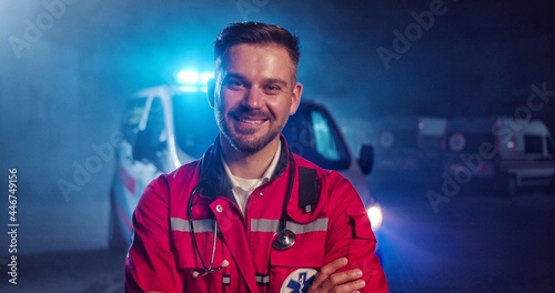 Portrait of Caucasian happy young male paramedic in red uniform smiling to camera and standing outdoor. Ambulance car on background. Attractive cherful male doctor at night shift. Call 911 photo